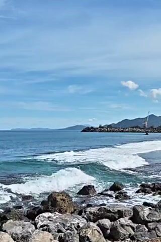 The video shows a scenic view of a rocky shoreline on a sunny day. Waves gently crash against the rocks in the foreground, creating white foam. The sea stretches out towards the horizon, where distant land is visible. A breakwater extends into the ocean, with construction cranes visible on it. The sky is blue with scattered white clouds. The overall scene is peaceful and serene.