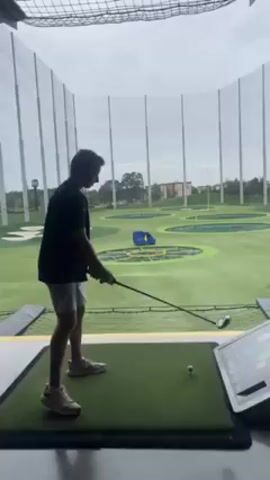 A person wearing a black shirt and light-colored shorts is playing golf at a Topgolf facility. The person is right-handed and takes a practice swing before hitting the golf ball. The target is a field with colored circles and targets, with a safety net visible in the background. A touchscreen console is to the person's right, and the environment appears to be an indoor golf bay. The sky is overcast.