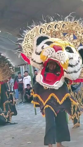 The video shows a street performance featuring a person in a large, elaborate costume resembling a tiger's head. The costume has a black and yellow striped head with red inside the mouth, large eyes, and a headdress adorned with feathers and gold accents. The body of the costume is black with yellow and red fringe details. Behind the performer, a crowd of spectators, including children and adults, are visible, some sitting and others standing. The performance appears to be taking place outdoors on a street or paved area. A large, circular object with red trim and a black surface with yellow lettering is also part of the performance.
