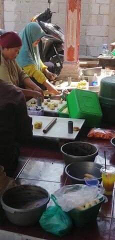 The video shows two women in an outdoor setting, likely a street market or food stall. The woman in the foreground, wearing a dark headscarf, is seated and appears to be working with some sort of dough. She is facing a tray filled with small, round items. To her left, another woman, wearing a light blue headscarf and a yellow shirt, is also seated and engaged in a similar activity, also working with dough. There are various containers and items spread around them, including plastic bags, buckets, and what appear to be food ingredients. A motorcycle is parked to the left of the frame. The ground is tiled. The scene suggests food preparation or selling.