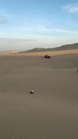 The video shows a desert landscape under a blue sky with scattered clouds. In the first shot, a person is kneeling on the sand while a red off-road vehicle is parked in the distance. The camera then pans to reveal a vast expanse of sand dunes. The next shot shows a group of three women standing on a dune, holding sandboards. In the final scene, two men, each carrying a sandboard, walk towards the women. The setting seems to be a recreational area where sandboarding is a common activity.