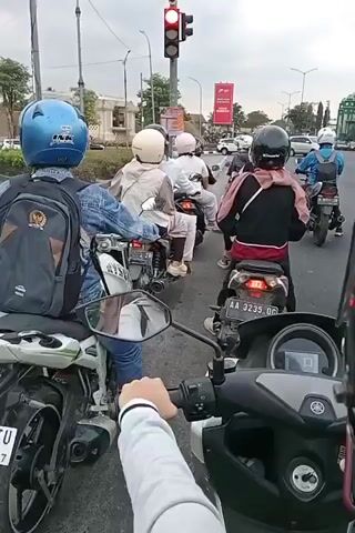 This video shows a first-person view of someone on a motorcycle waiting at a traffic light. The traffic light is red. Several other motorcycles are also waiting in front of and next to the camera. The riders on the other motorcycles are wearing helmets and some have passengers. The environment appears to be an urban street with buildings and trees visible in the background. The sky is overcast.