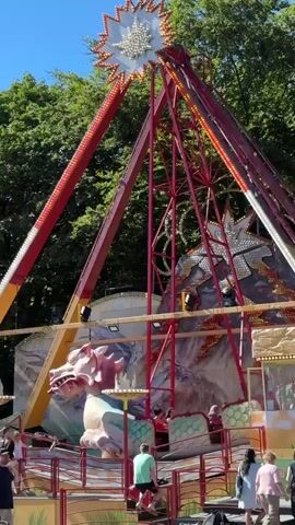 This media file captures a vibrant amusement park scene, likely during the daytime on a clear, sunny day, suggested by the bright lighting and blue sky. The primary subject is a large, elaborate amusement park ride that appears to be a pendulum swing or a pirate ship style ride. It features two towering, red, A-frame structures connected at the top by a spiky, starburst-like ornament adorned with lights. The ride itself has a large, dragon-themed gondola, with a detailed dragon head prominently displayed at the front. The backdrop behind the ride is painted with fantastical imagery, possibly mountains and other mythical elements, also featuring stars.

Several people are visible in the foreground and around the base of the ride, some appearing to be waiting to board or disembark, while others are simply present in the park. Their casual attire suggests a relaxed, recreational atmosphere. The setting is clearly outdoors, with lush green trees forming a natural backdrop behind the ride structures, indicating a park or fairground environment.

The overall mood conveyed is one of fun and excitement, typical of an amusement park experience. The bright colors of the ride, the people's clothing, and the sunny weather contribute to a cheerful and lively atmosphere. There is no visible text within the frame. The context provided places this scene in Taarbæk, Denmark.
