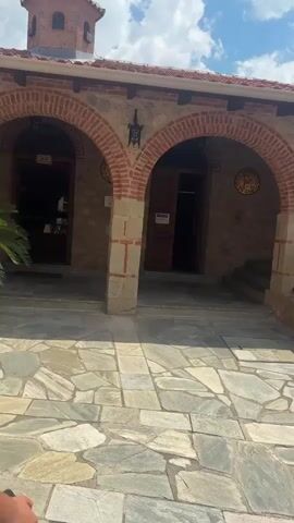 The video begins by showing an arched walkway with stone pillars and brick accents. The ground is paved with irregular stone slabs. A person wearing a black top and striped pants is visible walking down a set of stone stairs to the right. They are carrying a brown bag over their shoulder. The walkway leads to an entrance with a wooden door. Behind the walkway, a wooden railing is present, and beyond that, a lush green landscape can be seen, indicating an outdoor environment. The video continues to pan across the area, revealing more of the stone building with a red tiled roof and more arched walkways. In the background, a portion of a larger stone building with a wooden balcony and window is visible. Potted plants are placed within some of the arched alcoves. The sky is visible with scattered clouds.