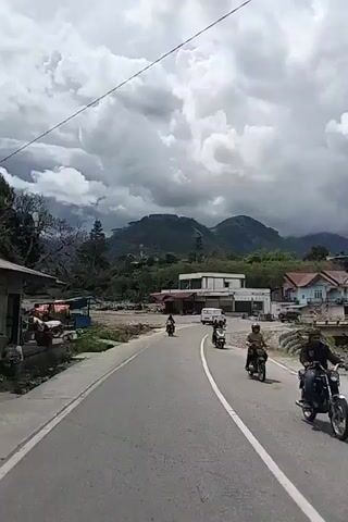 A motorcycle is traveling on a paved road. On the right side of the road, there is a group of people on another motorcycle. In the distance, there are buildings and mountains. The sky is cloudy.