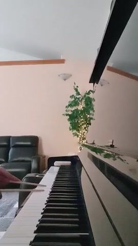 A close-up, low-angle shot of a person playing a piano. Their hands are visible on the keyboard, and drumsticks are held in their right hand, suggesting they are playing a rhythm. The piano is white with black keys, and the scene is indoors, with a dark leather couch and a potted plant with lights in the background. The lighting appears to be natural, coming from a ceiling fixture and possibly a window not visible in the frame. The overall impression is a casual, home-based musical performance.