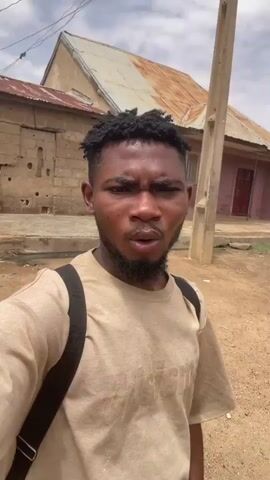 A young man is filming himself with his phone while walking down a dirt street. He is wearing a tan t-shirt, black shorts and sandals. He has a backpack on. The street is surrounded by houses with corrugated metal roofs. The sky is cloudy.