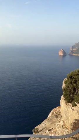 The video shows a vast expanse of deep blue ocean under a clear, pale blue sky. In the distance, a rocky island stands in the water, and beyond it, a mountainous coastline with rugged terrain and scattered vegetation. In the foreground, a stone cliff face is visible, topped with a bushy green shrub. A white railing follows the curve of the cliff, separating it from a paved viewing area. On this viewing area, two young men are present. One man, wearing a light blue t-shirt and dark shorts, is crouched down. The other man, shirtless with dark shorts, is standing and appears to be talking or gesturing. The camera is positioned at a high angle, looking down over the scene.
