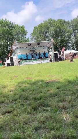 The video depicts an outdoor event on a sunny day in a large grassy field. A band is performing on a stage with a white backdrop on the left side of the field. Several individuals are seen walking on the grass near the stage and a portable toilet. To the right, multiple white and colored tents are set up, likely for vendors or stalls, with people moving among them. Further to the right, rows of wooden picnic tables and benches are arranged under tall trees. Numerous people are seated at these tables, some eating and drinking, while others are socializing. The sky is blue with some clouds, and a body of water is briefly visible in the background.