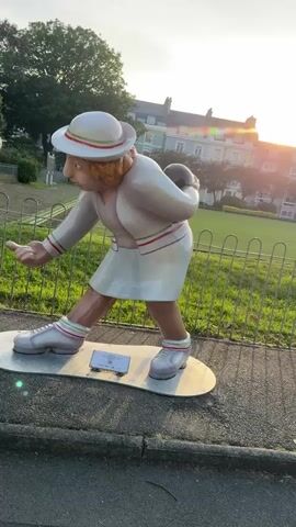 The video shows a painted wooden sculpture of a woman in a lawn bowling stance. She is wearing a white skirt and jacket with green and red trim, white sneakers with green and red socks, and a white hat with green and red trim. She is standing on a silver oval base, with a small black and white sign near her feet. She is holding a black lawn bowling ball behind her back and has her other arm extended as if about to release the ball. She has brown skin, a bulbous nose, and a stern expression. She is standing on a paved path next to a grassy lawn with a metal fence around it. In the background are trees and a light-colored building lit by the setting sun.