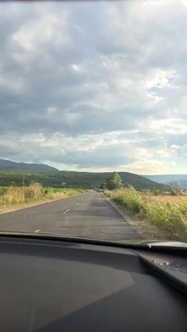 The video shows a view from a car driving on a paved road. The road has a dashed white line in the center. On either side of the road are grassy fields and green hills covered with trees. The sky is cloudy with patches of sunlight. Two cars and a van are visible in the distance, traveling in the same direction as the car from which the video is filmed.