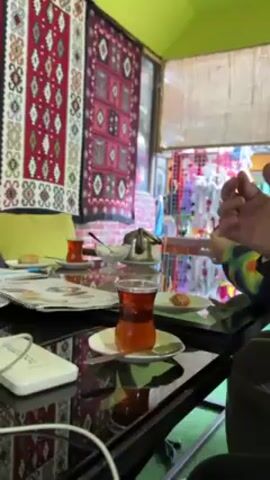 The video shows a table in what appears to be a cafe or restaurant. On the table are two glasses of tea, a small white dish with a dollop of something, perhaps yogurt, a small teapot, and two small plates with what looks like pastries on them. There is also a spiral-bound menu and a white, rectangular object, possibly a charger or power bank. A person's arm and part of their body are visible, and they reach to move a menu. The background features colorful rugs hanging on the walls and a window with colorful items displayed outside. The table is black and reflective. The room has a green ceiling.