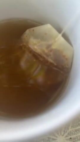 The video shows a close-up of a white mug filled with amber-colored liquid, likely tea. A tea bag, with its string visible, is submerged in the liquid. The tea bag appears to be releasing its contents, as evidenced by the swirling colors in the liquid and the slightly darker hue around the bag. The lighting is bright, highlighting the contents of the mug. There is no indication of any people or other objects in the immediate vicinity. The surface below the mug appears to be patterned with a light gray and white design. The video is shot from a top-down perspective, focusing on the process of the tea steeping.