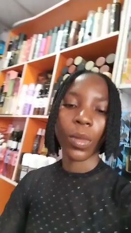 A young woman with braided hair and a black sheer top takes a selfie in what appears to be a store. She is positioned in front of shelves stocked with various perfume and cosmetic products. The shelves display numerous bottles and boxes of different brands and sizes. The lighting suggests an indoor environment, possibly a retail space. The woman smiles and tilts her head slightly as she records the video.
