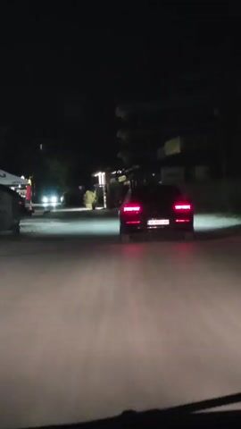 A black car with its brake lights illuminated is driving at night on a paved road. The car's license plate is visible and reads "B 4255 XB". In the background, there are some buildings and trees, with some lights visible from the buildings. The road appears to be illuminated by the car's headlights and possibly streetlights. The video is shaky, suggesting it was filmed from a moving vehicle.