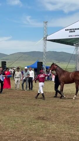 A horse race award ceremony is taking place outdoors. A brown horse with a red bridle is standing on a grassy field. A jockey in a red and white racing outfit is standing next to the horse, holding its reins. Several people are gathered around the horse, some wearing suits and formal attire, others in casual clothing. One person in a suit is holding a certificate or award. A large white tent with a logo is visible in the background, along with speakers and a stage structure. The sky is blue with some clouds, and hills are visible in the distance.