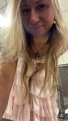 This media file features a woman with long blonde hair and a fair complexion, wearing a light pink, ruffled, layered dress. She is also wearing a delicate necklace with several charms. The woman appears to be in an indoor setting, possibly an airport terminal or a public waiting area, given the presence of seating with a metallic mesh back and overhead fluorescent lighting. The floor is tiled, and there are some vending machines or information kiosks in the background.

The woman is looking at the camera and smiling. She playfully twirls, causing her dress to swirl around her. The overall mood is lighthearted and joyful. She winks at the camera at one point. The scene appears to be taking place during the daytime, as indicated by the bright artificial lighting. The location is identified as Katunayake, Sri Lanka. There is no visible text in the media file.