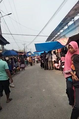 The video captures a bustling street market. A woman in a pink hijab and patterned skirt stands near a blue tarped stall. Several motorcycles are parked along the street, some with riders. Other people are visible walking and browsing among the stalls. The scene appears to be outdoors, with power lines overhead and a paved road surface. The lighting suggests it is daytime.