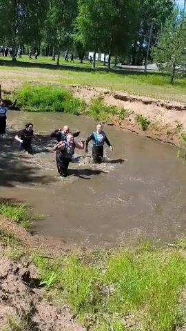 A group of people are wading through a muddy body of water. They are wearing athletic clothing, some with bib numbers. The water is shallow, reaching to their knees and lower thighs, and is filled with churning mud. The participants are moving forward, some with effort, as they navigate the obstacle. The environment appears to be an outdoor course with trees and greenery visible in the background, suggesting a race or challenging outdoor event.