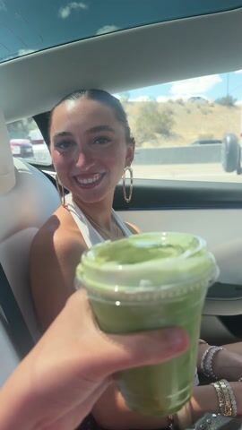 A young woman is sitting in the backseat of a car, wearing a white dress and hoop earrings. Someone hands her a green drink in a clear plastic cup with a lid. She smiles broadly at the camera. The car has white leather seats and a sunroof. Outside the car, a trailer and other vehicles are visible.