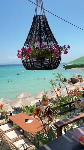 The video pans from a close-up of a hanging woven basket filled with pink flowers to reveal a picturesque beach scene. The beach has several straw umbrellas providing shade, and a number of people are visible swimming and relaxing in the clear turquoise water. The water transitions to a deeper blue in the distance, with faint landmasses on the horizon. In the foreground, a wooden table with wicker chairs is set up on a balcony or deck overlooking the beach. There are also potted plants and more flowers surrounding the seating area. The sky is a clear, bright blue, indicating a sunny day.