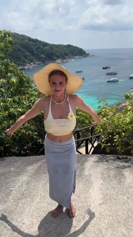 A woman in a straw hat, cream halter top, and light blue skirt stands on a stone platform overlooking a tropical ocean scene. She initially stands with arms outstretched, then puts her hands on her hat, poses while blowing a kiss to the camera, and dances with her hands. The background includes lush green vegetation, turquoise water with boats, and a partly cloudy sky.