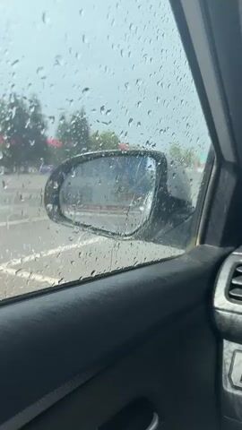 This is a first-person perspective video captured from inside a car, looking out the side window. The window is covered in raindrops, suggesting it is raining. The scene outside is a bit blurry due to the rain. Through the window and the side-view mirror, we can see a parking lot with white lane markings. In the background, there are trees and a building with a mural on its side. The mural depicts a figure in a blue and yellow costume, possibly a superhero or a character from a cartoon, with stars in the background. The building appears to be a school or similar public institution, indicated by the windows and architectural style. The sky is overcast, consistent with the rainy weather. The time of day appears to be daytime, likely afternoon or late morning, given the brightness of the sky. The overall mood is subdued, perhaps slightly melancholic, due to the rain and overcast conditions. The video does not contain any visible text. The location is identified as Verkhneyarkeyevo, Russia. The car's interior, including the door panel, is also visible in the foreground. The side-view mirror reflects the scene, providing a slightly different perspective of the building and the mural.