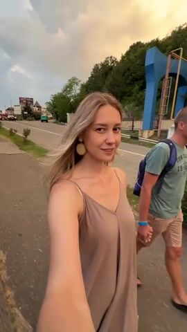 A blonde woman wearing a tan spaghetti strap dress and large circular earrings walks towards the camera, her hair blowing in the wind. Behind her, a man wearing a green t-shirt and khaki shorts, with a backpack, walks and looks towards the camera, smiling. They are walking on a paved path next to a road. In the background, there is a hill covered in green trees, a large blue structure, a few buildings, and a cloudy sky with a hint of orange. A car is seen driving on the road. The woman appears to be holding the camera, as it is positioned at her eye level.