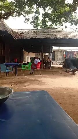 The video shows an outdoor scene in what appears to be a rural area. The foreground features a blue table, partially visible, with a metal bowl on it. The midground contains a simple structure with a corrugated iron roof, under which people are sitting on plastic chairs. Behind the structure, clothing is displayed for sale. In the background, cars drive by on a dirt road. A small fire is smoldering near the structure, and chickens are visible in the area. A large tree provides shade over the scene.