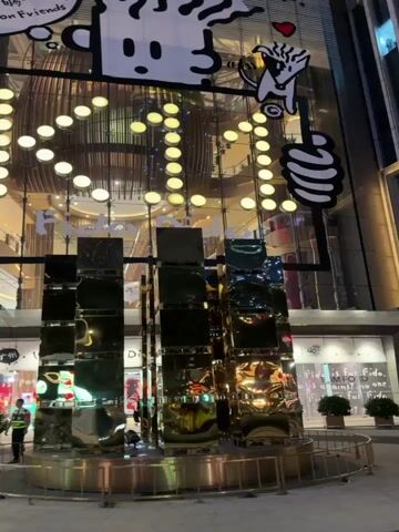 This media file captures a nighttime street scene in Tianhe District, China, likely a bustling commercial area. The setting is a modern urban environment with large, illuminated buildings.

The primary subject is the exterior of a multi-story building, showcasing a large digital display with the number "10" and a cartoon character at the top. Below this, a sign reads "Canton Friends." To the left of this display, the prominent facade of a GUCCI store is visible, featuring its logo and a large advertisement with a model. The architecture is contemporary, with illuminated geometric patterns on the GUCCI building and vertical golden lights on another section of the structure.

Several people are present in the scene, walking on the paved ground. Some are casually dressed, while others appear to be shoppers or passersby. A security guard in uniform is also visible near the "Canton Friends" sign. The overall atmosphere suggests a lively evening in a shopping district.

Notable details include the time of day, indicated by the artificial lighting and the darkness of the sky. The presence of reflections on the building's facade and on what appears to be a water feature or sculpture in the foreground suggests a clear night. The overall impression is one of a vibrant, well-lit, and affluent urban area.