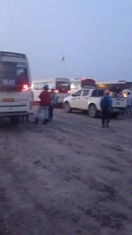 A group of people are standing around in a dusty, outdoor environment. There are several vehicles present, including a white pickup truck with "SAFETY PATROL" written on the side, and other trucks and buses in the background. The sky is overcast and the lighting suggests it may be early morning or late afternoon. Some of the people are wearing hard hats and high-visibility vests, indicating they are likely workers on a construction site or similar industrial area. One person is kneeling on the ground. The ground is rough and uneven, with tire tracks visible.