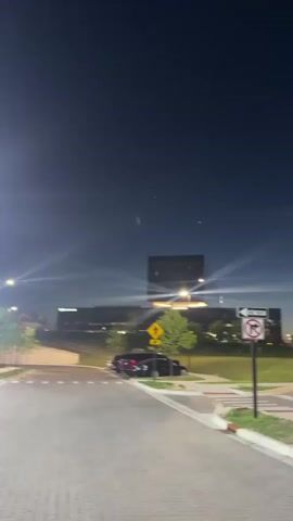 The video shows a nighttime scene in what appears to be an urban or suburban area. The camera pans slowly from left to right, revealing a street or pathway paved with cobblestones on the left, transitioning to an asphalt parking lot on the right.

The sky is dark blue, indicative of night, with several bright specks that could be stars or distant lights. Bright streetlights create prominent starburst effects in the frame. In the background, large buildings are visible, some with illuminated windows.

In the foreground, there are small trees planted in grassy areas alongside the paving. Several cars are parked in the lot, including a blue Jeep Cherokee and a black sedan, among others.

Two street signs are visible: a yellow diamond-shaped sign with a black arrow indicating a curve to the right, and a white rectangular sign with a red circle and a diagonal line through a right-turn arrow, below which the text "ONE WAY" is clearly legible. No people are visible in the video. The overall atmosphere is quiet, typical of an evening in a commercial or residential complex.
