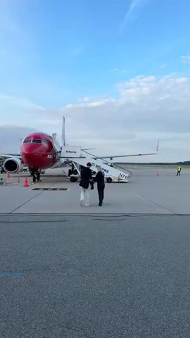 Two individuals, one in white pants and a dark jacket and the other in dark clothing, walk up a mobile staircase towards an airplane. The airplane is red and white with "norwegian" visible on its side. The staircase is attached to the plane's door. The scene takes place on an airport tarmac with the sky visible in the background.