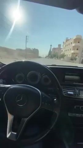 The video shows the interior of a Mercedes-Benz car, likely a model from the early 2010s based on the dashboard design, from the driver's perspective. The car is driving down a road in a Middle Eastern country with houses and a mosque visible in the background. The sky is clear and sunny. The video briefly shows the driver in the rearview mirror.