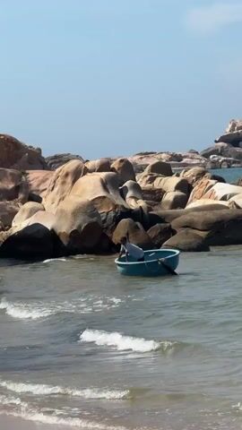 A person in a blue boat rows through the water near a rocky shoreline. A small wooden structure on stilts is visible on the left side of the frame. The water is clear and calm, with small waves breaking on the sandy shore. The sky is clear and blue.