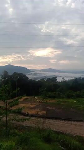 The video captures a sunrise over a misty, mountainous landscape. In the foreground, there is a dirt clearing with some construction equipment visible, including a large electrical tower. Lush green trees and foliage dominate the midground and background. The sky is partly cloudy, with the sun breaking through the clouds, casting a warm glow. The overall atmosphere is serene and picturesque, with the mist adding a touch of mystery to the scene.