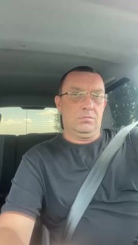 A middle-aged Caucasian man with short, dark hair and glasses is sitting in the driver's seat of a car. He is wearing a black t-shirt and a seatbelt. His right fist is clenched and raised, and he is looking to his right. The interior of the car is visible, including the headrest of the passenger seat and the headliner. The background outside the car appears to be a bright, sunny day with some trees visible. The man's expression is serious.