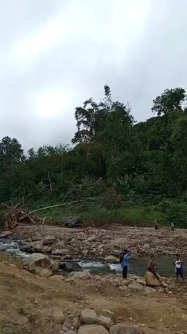 The video shows a group of people gathered near a rocky riverbed under a cloudy sky. Lush green trees cover the banks and the background. Several individuals are visible: some are standing by the water's edge, while others are walking along the rocky terrain. One person is seated on a boulder. A small truck and some debris are seen further along the bank. The water flows through a small constructed weir.