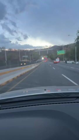 The video shows a view from the driver's seat of a car traveling on a multi-lane highway under a cloudy sky. Other vehicles are visible ahead on the road, including cars, trucks, and what appears to be a work vehicle with flashing lights. A bridge is visible in the distance. There are roadside barriers on the left and signs on the right.  The dashboard of the car is visible in the foreground.