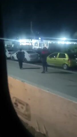 At night, a man in dark clothing walks on a street. Another man in a red shirt stands further down the road. Cars are parked on the side of the street, and a gas station is visible in the background, brightly lit. A concrete barrier runs along the bottom of the frame. The video is taken from inside a vehicle. The man in dark clothing adjusts his head covering as he walks.