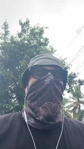 The video shows a person wearing a black t-shirt, a black bucket hat, sunglasses, and a patterned neck gaiter that covers their nose and mouth. White wired earbuds are visible. The person appears to be walking outdoors, with trees and palm trees in the background, along with some power lines against a cloudy sky. The camera is positioned at eye level and is slightly tilted upwards. The person's face is obscured by the accessories. The overall scene suggests an outdoor activity, possibly a walk or jog, in a tropical or subtropical environment.