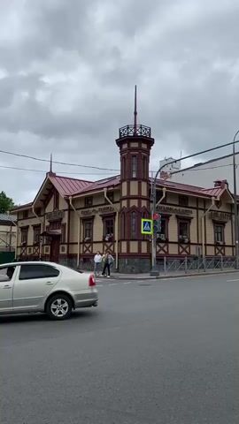 A street view shows a charming, historic wooden building with a red roof and intricate trim in shades of burgundy and cream. The building features a distinctive turret with a spire, several windows with flower boxes, and signage in Cyrillic script. The street in front of the building is paved, with a pedestrian crossing marked by a yellow sign. Two people, casually dressed, stand near the corner of the building, seemingly engaged in conversation. A dark-colored SUV drives past the intersection, and the sky is overcast with a cloudy, gray appearance. The overall impression is one of a quiet, overcast day in a town with well-preserved architecture.