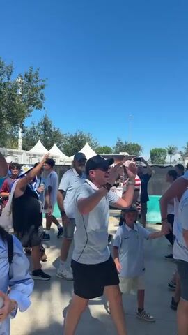 The video shows a crowd of people walking outdoors on a sunny day. Many people are wearing blue and white clothing, and some are holding flags. In the foreground, a woman in a light blue shirt and sunglasses is holding a bottle of water. Behind her, a man in a light blue shirt and black shorts walks by. In the background, there are tents, trees, and a large building that appears to be a stadium. Some people are sitting on the grass. The atmosphere appears festive and energetic, likely indicating an event or gathering.