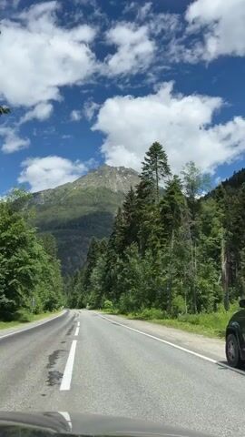 A scenic view of a winding road through a dense forest, leading towards a majestic mountain under a bright blue sky dotted with fluffy white clouds. The road is paved with asphalt and has white lane markings. Tall evergreen trees line both sides of the road, creating a tunnel-like effect. The mountain in the distance is covered in lush green vegetation, with patches of rock visible on its slopes. The sun is shining, casting shadows on the road and highlighting the vibrant green of the trees. The overall impression is one of a peaceful and beautiful journey through nature.