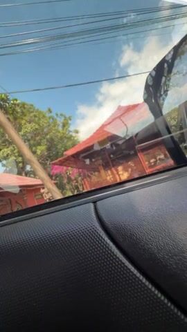 The video begins inside a vehicle, showing a view through the windshield and side window. The exterior scene is blurred but shows a red-roofed structure with a cage-like enclosure. A young boy in a brown shirt and patterned sarong stands in front of the structure, holding a stick. He turns to face the camera as the vehicle moves closer. The perspective shifts to eye-level with the boy as the video continues, showing him standing on a paved area next to a curb. The red-roofed structure, which appears to be a shrine or temple with people inside, is visible in the background. The boy holds the stick with both hands. The camera then moves away from the boy, and the scene returns to a view from inside the vehicle, showing the boy and the structure again.