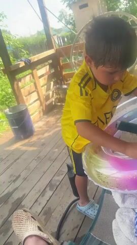 The video opens with a close-up shot of a person's foot wearing a tan shoe with holes. The camera then pans up to show a portion of their leg, which has several red marks, possibly insect bites. A child in a yellow t-shirt and black shorts is visible in the background, holding a colorful inflatable ring. They are standing on a wooden deck. The setting appears to be outdoors, with greenery and a wooden structure in the background. The child is facing away from the camera and seems to be engaged in an activity with the inflatable ring.