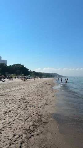 This video showcases a bright, sunny day at Timmendorfer Strand, Germany. The scene is a sandy beach with clear blue water gently lapping the shore. Several people are visible enjoying the beach and the sea. Some are wading in the shallow water, while others appear to be swimming further out. In the background, a line of trees and some buildings suggest a developed coastline. The sky is a vibrant, cloudless blue, indicating fair weather. The time of day appears to be midday or early afternoon, given the bright sunlight and the activity on the beach. The overall mood is relaxed and leisurely, typical of a summer day at the seaside. No text is visible in the media.