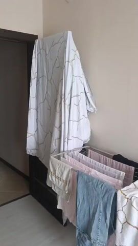 A drying rack is positioned next to a dark dresser, partially covered by a white sheet with a cracked gold pattern. Several items of clothing, including jeans and shirts, are draped over the drying rack. Another white sheet with the same pattern is draped over the other end of the rack. To the right, a stack of folded clothes is visible. The floor is light-colored and appears to be wood or laminate.