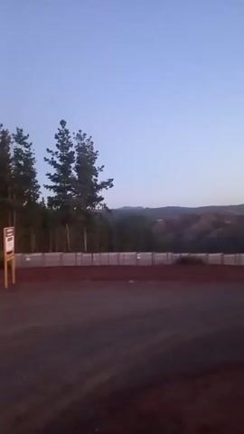 The video depicts a scene with a dark, dirt-covered ground in the foreground. Behind it, there is a long, concrete barrier fence. Beyond the fence, a line of trees is visible, and in the far distance, there are rolling hills. The sky is a pale blue color. A yellow post stands on the right side of the frame. The video appears to be taken in the early morning or late evening due to the dim lighting.