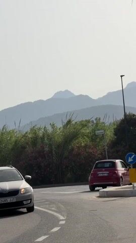 A video shows a street scene with cars and people. In the foreground, a silver car is driving on a road. In the background, there are mountains and lush green foliage. A red car is parked on the right side of the road, near a traffic sign. A white van with "AmicoBlu" written on it is also visible, and two people are crossing the street in front of it. The sky is hazy.